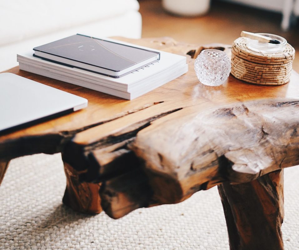 Massiver, mittelbrauner Couchtisch aus Holz steht auf einem weißen Teppich.Auf dem Tisch liegen Bücher und ein Laptop sowie ein Windlicht aus Glas.