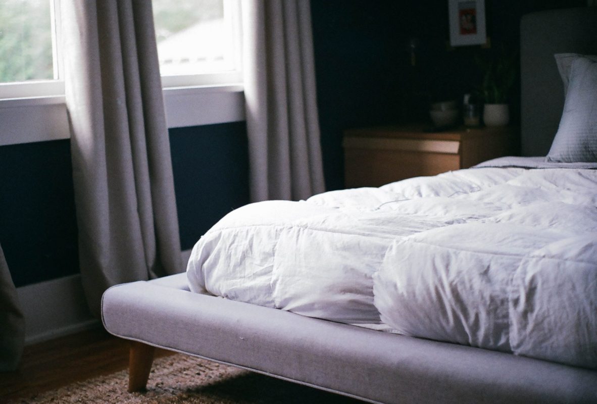 Schlafzimmer mit hellgrauem Stoffbett mit weißer Mattratze. Die zwei Fenster zieren zwei bodenlange, graue Vorhänge.